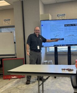Crane Tech instructor Dan Calabro teaching a training class while standing beside a large digital display showing hook inspection information and equipment safety guidelines.